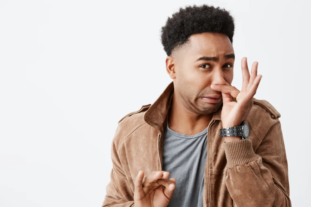 That smells really bad. Close up portrait of young funny dark-skinned man with afro hairstyle closing nose with fingers, feeling ad from disgusting smell from rubbish dump in city. Copy space.