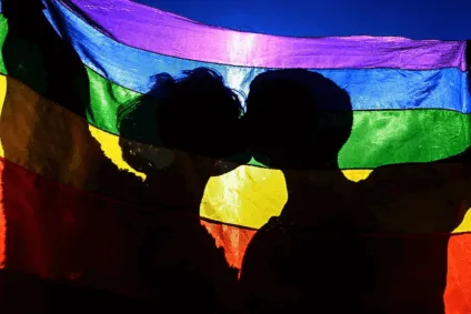 Dois jovens se beijam sob a bandeira LGBTI+, simbolizando o orgulho LGBTI+ como resistência e afeto em um país que ainda nega direitos e dignidade.